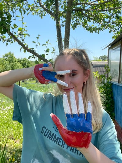 A young blonde woman stands outdoors in a grassy area with a tree overhead and a building in the background, likely Yasnoe, Russia. She is wearing a teal t-shirt with dark lettering that reads "LE SUMMER PARADISE." Her right hand is painted with the colors of the Russian flag: red on the palm and lower fingers, and blue on the upper fingers. Her left hand is also painted with red and blue on the palm and lower fingers, but the upper fingers are painted white. She is holding her hands up to her face, with the fingers of her left hand extended and partially obscuring her eyes and nose. Her right hand is held closer to her chest. The background includes green grass, white wildflowers, trees, and a blue wooden structure with glass panels. The sky is clear and blue.