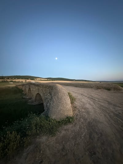 This is a nighttime photo of an ancient Roman aqueduct in Filipeni, Moldova. The aqueduct, made of stone, spans across a grassy field. The moon is visible in the clear, blue night sky, suggesting it is a full moon. The overall mood of the image is serene and peaceful. There is no visible text in the image.