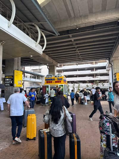 People stand with their luggage outside a building under an overhang. A yellow and blue booth advertising "Minibus Taxi" in multiple languages is prominent. Signs with arrows and building directory are visible. Travelers are diverse, carrying suitcases of various colors and designs. The scene suggests a transportation hub like an airport or bus station.