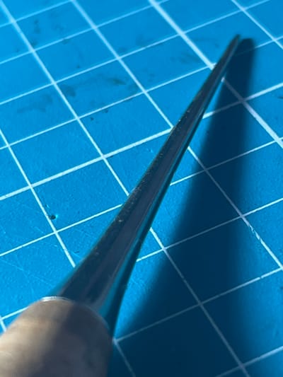 The image shows a close-up, high-angle view of a pointed tool, possibly an awl or a scribe, lying on a blue grid cutting mat. The tool has a wooden handle and a metallic, tapered point. The cutting mat has white grid lines, and some dark specks are visible on the mat. The lighting creates a shadow of the tool on the mat. There is no visible indication of the city of Novy Urengoy, Russia, or any people. The scene appears to be a still-life arrangement of a crafting or repair tool.
