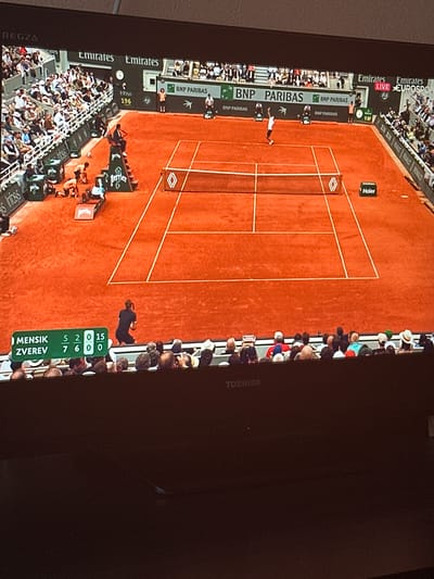 This media file is a photograph taken in Marikostinovo, Bulgaria, depicting a live tennis match broadcast on a television screen, rotated into a portrait orientation.
The image prominently features a bright red clay tennis court with white lines, typical of tournaments like the French Open. A tennis player, dressed in white, is visible mid-action near the center of the court. Spectators fill the surrounding stands, indicating a professional event.
Visible on-screen are numerous sponsor logos, including "Emirates," "EUROSPORT," "BNP PARIBAS," "Haier," "Perrier," "Infosys," and "RENAULT." A scoreboard clearly displays player names and scores: "MENSIK" (Jakub Mensik) and "ZVEREV" (Alexander Zverev). The score indicates Zverev won the first two sets (7-5, 6-2), and the current game in the third set stands at 0-0, with Mensik having 15 points and Zverev 0. "LIVE EUROSPORT" is visible, confirming a live broadcast. Other numerical indicators "0:37" and "196" are also shown on the broadcast overlay.
The match is taking place during the daytime under sunny conditions, casting distinct shadows on the court. The television screen itself displays "REGZA" on the bottom left and "TOSHIBA" on the right frame, identifying the display device.