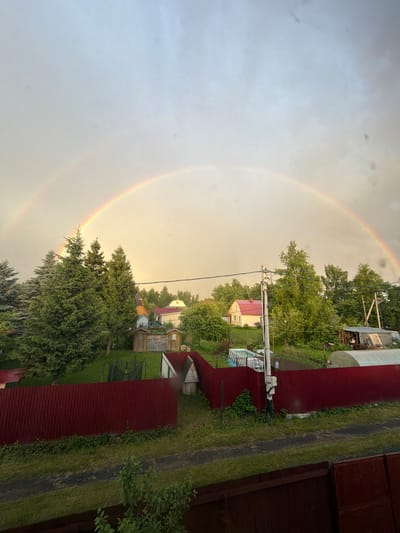 This image captures a rural landscape in Манихино Снт, Russia, under a sky transitioning from grey to a warm, pale yellow, indicative of the aftermath of rain or the approach of evening. The most striking element is a vibrant double rainbow arcing across the sky, its colors soft but discernible against the diffused light.
The scene is dominated by lush green trees, dense and varied in their foliage, interspersed with small buildings and structures characteristic of a dacha or summer home community. Red corrugated metal roofs are visible on several buildings, adding a bold visual contrast to the greenery. Among the structures, we can see what appear to be sheds, a greenhouse with a white frame, and a house with a red roof and yellow walls. In the foreground, on the right side of the image, a dark wooden fence runs along the edge of the property. A well-maintained grassy area and a path or dirt track are also present.
The time of day is likely late afternoon or early evening, given the soft, diffused light and the presence of the rainbow. The weather has recently been rainy, which has contributed to the vividness of the rainbow and the lushness of the vegetation. There is no visible text in the image. The overall impression is one of peaceful, natural beauty, with the rainbow adding a touch of magic to the serene rural setting.
