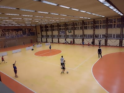 An elevated view captures an indoor sports gymnasium in Fedorovka, Russia. The facility features a light orange and orange two-toned synthetic court with white demarcation lines, including a central oval and a large curved boundary on the right. The high ceiling is fitted with numerous horizontal fluorescent light fixtures. The upper walls consist of a series of windows, while the lower sections are paneled in beige with a dark patterned trim. A large mural depicting athletes is visible on the far left wall, beneath which a dark sports goal is positioned. A digital scoreboard displaying "0:04" and other data is mounted on the far wall. Approximately 20 individuals, predominantly young males, are present on the court. Most are dressed in athletic attire (t-shirts, shorts) and are engaged in activities consistent with soccer or futsal training, with several handling soccer balls. Multiple white, low barriers or small goals are deployed in the mid-ground. An adult male in dark athletic wear stands on the right side of the orange court area, observing the activity.