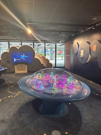 The image captures an indoor scene within what appears to be a modern science museum or exhibition hall, likely the Experimentarium in Copenhagen, given the visible Danish text and location context.
In the center, a prominent exhibit features a large, clear, oval-shaped dome resting on a dark blue, circular base. Inside the dome, numerous U-shaped elements glow vibrantly in neon colors like pink, purple, blue, and green, suggesting a dynamic light display or an interactive scientific demonstration. The floor in the foreground is covered with a dark grey carpet adorned with a pattern of scattered yellow and light grey circles.
To the left, a large, dark grey, cloud-shaped structure houses a screen displaying an animation of the Earth in space with colorful splatters and the word "TAKEOFF" at the bottom, indicating another interactive or informational exhibit.
Along the dark wall in the background, several circular display panels are mounted, showcasing diagrams, illustrations, and descriptive text. One panel clearly states "SÅDAN GØR DU DIG KLAR" (Danish for "HOW TO PREPARE"), and another partially visible panel includes "EXPERIMENTARIUM" and "TIDEN?" (Danish for "THE TIME?"), confirming the venue's identity.
Large windows on the far side offer a view of an outdoor urban setting with buildings, one of which has "SIX CLASS TOWN" visible on its facade. The interior is well-lit by both natural light from the windows and artificial spotlights from the paneled ceiling. The scene appears to be set during daytime. No people are present in the frame.