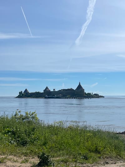 In the city of Sheremet'evka, Russia, a fortified island fortress stands on a body of water, likely a river or lake, under a bright, clear sky. Two distinct jet trails are visible, one a long, straight line from upper left to lower right, and the other a more wispy, curved trail in the upper right quadrant. The fortress, constructed of dark stone, features several conical towers and a long, fortified wall. A wooden pier extends from the fortress into the water, with a few small boats docked. The foreground of the image is dominated by a lush, green embankment covered in tall grass and some scattered bushes. The water is calm and reflects the blue of the sky.