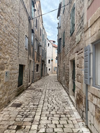 This is a ground-level, eye-level, medium shot of a narrow cobblestone alleyway in the historic town of Stari Grad, Croatia. The alley is flanked by tall, ancient stone buildings. The stone construction is visible on both sides of the alley, with rough-hewn blocks forming the walls. Many windows are present, some with weathered, louvered shutters, predominantly in shades of green and grey. The cobblestone pavement is uneven and shows signs of age and wear, with some sections appearing damp or wet. There are various architectural details, including doorways, drainpipes, and electrical conduits running along the building facades. A street lamp is visible in the distance, illuminating a portion of the alley. The sky above is overcast and cloudy. No people are present in the scene. The overall impression is one of a well-preserved, historic urban environment.