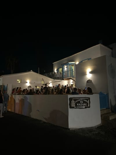 The image depicts the exterior of "To Kazani," a family tavern located in Aradippou, Larnaca District, Cyprus, at night. The scene is illuminated by multiple wall-mounted lights, casting shadows. Two white-painted buildings are visible: a main two-story structure featuring a balcony with blue railings and a blue-framed door/window on the upper level, and a smaller, single-story building to its left.

Approximately 30-40 individuals, primarily adults, are gathered behind a low, white retaining wall that forms the foreground barrier. Most are standing and appear to be socializing. A few individuals are also visible on the upper balcony of the two-story building. One person on the far right of the crowd is holding a smartphone in an upright position.

A dark, rectangular sign with white lettering is affixed to the prominent white wall in the foreground, reading "ΟΙΚΟΓΕΝΕΙΑΚΗ ΤΑΒΕΡΝΑ ΤΟ KAZANI" (Family Tavern To Kazani) and "ΤΗΛ 99 31 32 30". The ground in the immediate foreground is dark and paved. A segment of a blue wooden fence is visible on the far right. A palm tree is partially visible on the left side of the frame, behind the single-story building.