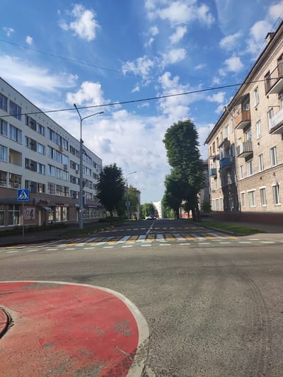 The image depicts a street scene in Polotsk, Belarus, on a clear day with scattered white clouds in a blue sky. A pedestrian crossing with yellow and white stripes is visible in the foreground. On the left side of the street, a multi-story residential building with a light-colored facade and numerous windows is present. A blue pedestrian crossing sign with a white silhouette of a person is also visible on this side. Further down the street, trees line the median. On the right side of the street, a multi-story brick building with balconies is shown. The street is paved and appears to be an urban environment. A red painted circular traffic island is partially visible in the lower left corner of the image. The scene appears to be captured during daylight hours.