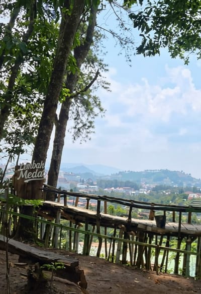 In Lembang, Indonesia, a scenic overlook offers a view of rolling hills and a town nestled in the valley. A wooden sign on a tree trunk reads "Lembah Meda". A rustic wooden walkway, elevated by bamboo stilts, extends towards the vista, with a dark bucket resting on its surface. In the foreground, a rough-hewn wooden bench sits on a dirt path, partially shaded by the overhanging leaves of lush trees. The sky above is a bright blue, dotted with fluffy white clouds.