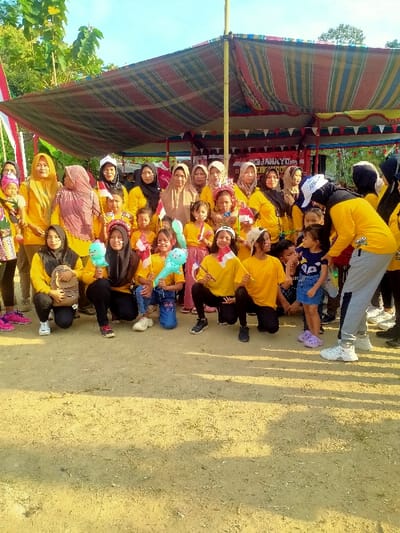The image displays an outdoor group portrait of approximately 35 individuals, predominantly adult women and children, located in Jetis, Indonesia. The subjects are positioned on a light brown, dry earth surface beneath a large, horizontally striped fabric canopy supported by multiple poles. The canopy features red, blue, yellow, and green stripes.
The majority of individuals are wearing yellow tops, with several adult women wearing headscarves in colors such as brown, black, and cream. In the foreground, several children and young adults are kneeling or crouching. Multiple children are holding small Indonesian flags, which are red and white. Two young women in the kneeling row hold light blue, elongated balloons shaped like cartoon characters. On the far right, an adult female wearing a yellow shirt, grey sweatpants, and a white baseball cap is bent at the waist, facing and interacting with a small child in a blue denim skirt.
In the background, beneath the canopy, a red banner with partially legible white text, including the letters "JAHA", is visible. Beyond the canopy, green foliage and trees are visible against a clear blue sky, indicating an open-air environment. The scene is illuminated by natural daylight, casting subtle shadows on the ground.