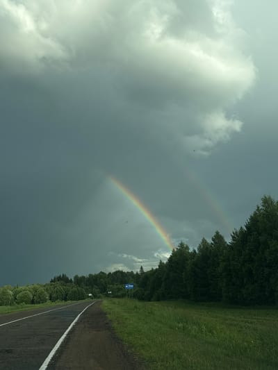 A double rainbow is visible in a dark, cloudy sky over a rural landscape in Кватчи, Russia. A paved road with a white line marking the shoulder runs from the lower left towards the distance. Lush green trees line the right side of the road, extending into a dense forest. The ground on the right is covered in green grass and some wildflowers. A blue road sign with a Gazprom logo and an arrow pointing to the left is visible near the edge of the trees. The sky is heavily clouded with a mix of dark storm clouds and lighter, brighter areas where the sun is breaking through, illuminating the rainbows.