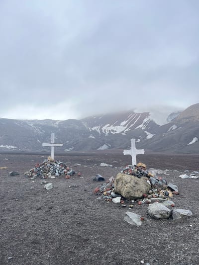 Two white crosses stand on a dark, rocky terrain. Each is surrounded by small piles of colorful stones. In the background, a steep, snow-streaked mountain rises, partially obscured by low-hanging clouds or fog. The sky is overcast, creating a somber atmosphere. The scene is tilted to the side, giving it an unsettling perspective.