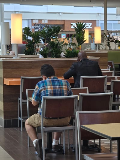 This image depicts an indoor scene, likely a food court or common area in a shopping mall, with a focus on two individuals seated at tables. The setting features wooden paneling, metal furniture, and large, rectangular light fixtures. Green plants are visible, adding a touch of nature to the otherwise artificial environment.

The primary subjects are two men. One man, facing away from the camera, is wearing a black textured shirt and appears to be bald. He is seated at a table with a dark wooden top. The second man, also facing away, is wearing a blue and orange plaid shirt and khaki shorts. He is seated at a table with a dark wooden top and is leaning forward, with his arms resting on the table. His legs are visible, showing him wearing white socks and brown shoes.

The activity appears to be casual dining or relaxation. The overall mood is subdued, with the subjects seemingly engaged in their own activities. The lighting suggests it is daytime, but no specific weather conditions can be inferred from the indoor setting.

Visible text includes "SUBWAY" on a sign in the background, indicating a potential dining establishment. Other signs are present but illegible. The presence of mall-like architecture and the "SUBWAY" sign strongly suggest a location within a shopping center. The provided location context of Alpharetta, United States of America, further refines the setting.