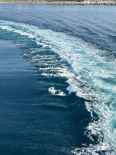 The image displays a maritime scene near Kamariotissa, Greece, captured from an elevated perspective, likely a moving vessel. The majority of the frame is dominated by deep blue ocean water. A prominent wake, generated by a vessel, extends diagonally from the upper left to the lower right, characterized by turbulent, aerated water appearing white and light blue. This frothy wake sharply contrasts with the calmer, rippled dark blue water on both sides. In the distant background, a strip of coastline is visible, featuring a light-colored linear structure, possibly a road or seawall, bordered by sparse vegetation and indistinct man-made buildings. No human figures or specific identifiable objects are present in the immediate marine environment.
