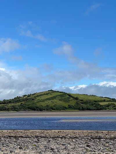 This is a landscape photograph taken at Dunuisce, Ireland. The image is oriented vertically. On the left, a vast expanse of bright blue sky is dotted with white and gray clouds. The sky occupies roughly the left half of the frame.
The right half of the image depicts a coastal scene. A green, gently sloping hillside covered in dense vegetation dominates the upper right portion. The hillside appears to be a pasture or moorland. Below the hillside is a strip of light-colored sand or mudflats, indicative of a tidal area. Further to the right, a body of blue water with subtle ripples stretches into the distance. The foreground on the far right is a rough, rocky shore, with many small to medium-sized stones and some scattered seaweed. The overall impression is a bright, sunny day with scattered clouds, likely sometime during the day. There are no people or man-made objects clearly visible, only natural elements. The terrain suggests a scenic, undeveloped coastal location.