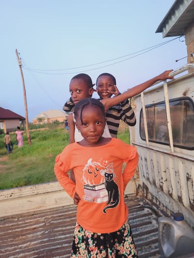 Three children are positioned within the open bed of a white utility vehicle or pickup truck, located in the city of Shen, Nigeria. The foreground is occupied by a young girl with dark skin and braided hair, wearing an orange long-sleeved shirt with a black cat, pumpkin, and book motif, and a black floral skirt. She stands centrally with her hands on her hips, looking towards the camera with a neutral expression. Behind her, slightly to the viewer's left, stands a boy with dark skin and short hair, wearing a white top with dark horizontal stripes, also looking towards the camera. Further back and to the viewer's right, a third child, a boy with dark skin and short hair, smiles broadly and displays a peace sign with his left hand, which rests on a white metal railing of the vehicle. He wears a striped shirt. The truck bed features corrugated metal flooring and worn white metal railings; a grey plastic container with a blue cap is visible on the bed's floor in the lower right quadrant. The background consists of a clear, light blue sky, green vegetation, a leaning wooden utility pole on the far left, and multiple single-story residential buildings with varying roof colors in the mid-distance. The scene is illuminated by natural daylight.
