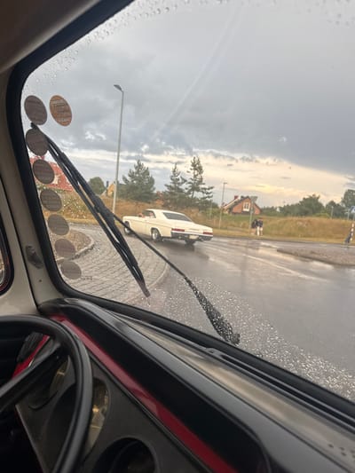 The image is taken from inside a vehicle looking out through a rain-streaked windshield. A white vintage car is driving away from the camera on a wet road. There are two people walking in the distance on the sidewalk. The environment appears to be a residential area with houses and trees visible. The sky is cloudy, suggesting inclement weather. There are several circular stickers on the lower left portion of the windshield, which appear to be vehicle inspection stickers. The car interior, including a steering wheel and dashboard, is visible in the foreground. The location is identified as Falkenberg, Sweden.