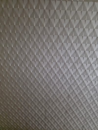 The image is a close-up, angled view of a textured, grey, geometric wall covering. The texture consists of a repeating pattern of small, raised pyramids or faceted shapes. The lighting creates shadows and highlights that emphasize the three-dimensional nature of the surface. There are no people, objects, or actions visible in the image. The image does not provide any information about its location in Valga city, Estonia.
