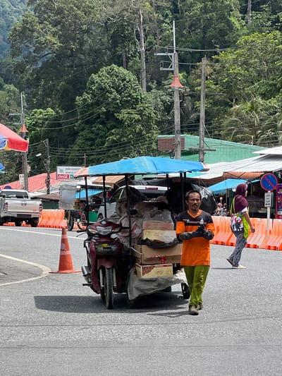 This media file depicts a street scene in Tambon Wang Prachan, Thailand. The primary subjects are two individuals, likely a vendor and a customer, interacting near a cart laden with goods. The vendor, a man wearing an orange shirt and green pants, stands next to the cart. He is wearing dark gloves and appears to be holding something. The cart itself is covered with a blue tarp and is filled with boxes and bags, suggesting it is a mobile stall or a delivery vehicle. In the background, another person, possibly a customer, is visible wearing a purple headscarf and a colorful shirt, standing behind orange traffic barriers.
The scene appears to be set in a bustling outdoor market or roadside. The presence of overhead power lines, lush green trees in the background, and the colorful awnings of nearby stalls indicate a tropical environment. The weather seems fair, with bright sunlight casting shadows on the asphalt road, suggesting it is daytime. The overall atmosphere seems to be that of commerce and daily life, with people engaged in transactions or preparing for them.
Visible text includes a Coca-Cola logo on a sign attached to one of the stalls. Other signs are present but too blurry to read. The road has painted white lines, and orange traffic cones are placed along the side, possibly indicating a temporary closure or a designated area. The presence of a red motorcycle attached to the cart suggests it is the mode of transport for the vendor's goods.