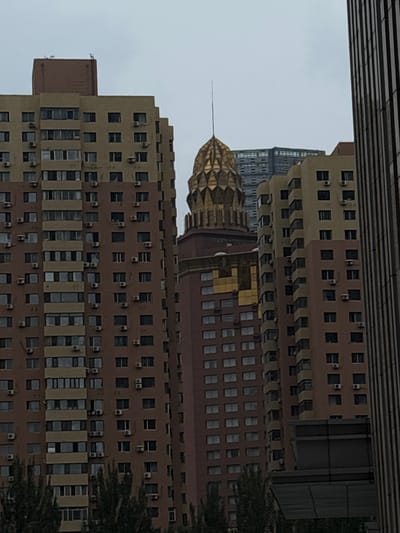 In Heping District, China, a cluster of apartment buildings and a distinct gold-domed structure are visible under an overcast sky. The buildings are primarily residential, characterized by numerous windows and external air conditioning units. One of the buildings on the left is a tall, light brown and tan structure with many rows of windows and AC units. Adjacent to it, on the right, is a similar, though slightly narrower, building in shades of tan and pink. Between these two, a striking building with a unique gold, faceted dome topped with a slender spire stands out. Further in the background, another modern, glass-fronted building is partially obscured. The foreground and lower portions of the buildings are lined with dark green trees. The overall scene suggests an urban environment with a mix of architectural styles.