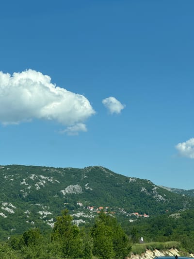 The image shows a mountainous landscape with a village nestled in the valley. The sky is a clear blue with a few white clouds. The mountains are covered in green vegetation, with some exposed rocky areas. The village has several houses with red tile roofs. In the foreground, there are some trees and bushes. A portion of a dark-colored vehicle is visible in the lower right corner. This scene is located in the vicinity of Obzovica, Montenegro.