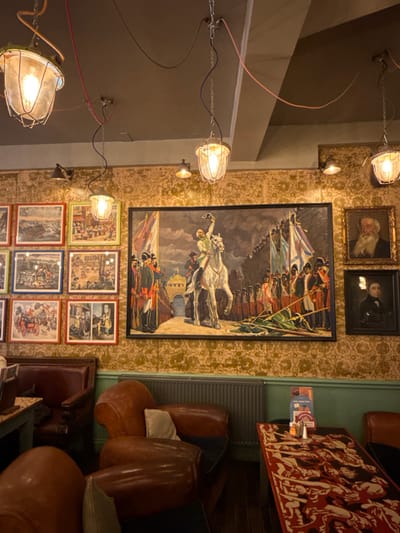 This image depicts the interior of The Feathers Inn, located on Beacon Street in Lichfield, United Kingdom. The scene is a dimly lit, unoccupied seating area characterized by historical and ornate decor.
The upper walls are covered in gold-toned wallpaper featuring a repeating damask pattern, transitioning to a muted teal or sage green lower wall section. The dark grey ceiling features exposed electrical wiring in red, blue, and green, from which multiple industrial-style pendant lights with exposed bulbs encased in clear glass and metal cages hang via chains. Additional wall-mounted spotlights cast warm illumination.
The central focal point on the far wall is a large, framed painting or tapestry depicting a historical military scene. It prominently features a figure resembling Napoleon Bonaparte on a white horse, raising his right arm, surrounded by soldiers and flags, with an archway visible in the background. To the left, a gallery wall displays approximately ten smaller framed prints, each in a distinct colorful frame (red, green, blue, brown), illustrating various historical or scenic subjects. To the right of the large artwork, two vertically stacked framed portraits depict an elderly bearded man (upper) and a younger man in dark attire (lower).
Seating arrangements include several distressed brown leather armchairs and parts of sofas. In the foreground, two such armchairs are visible, one featuring a small, light-colored cushion. A dark wooden table in the right foreground is adorned with a distinctive red and black patterned runner depicting stylized human figures. A small metal salt/pepper shaker and a menu holder are present on this table. A grey, ribbed radiator is affixed to the lower green wall beneath the central painting. The flooring appears to be dark wood. No individuals are present in the scene.