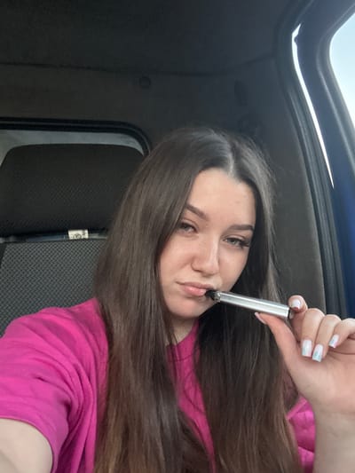 A young woman with long, dark brown hair and fair skin is pictured indoors, holding a silver vape pen to her lips. She is wearing a bright pink t-shirt. The background appears to be the interior of a vehicle, with a sunroof visible at the top of the frame. The lighting suggests it is daytime, possibly with bright sunlight coming through the sunroof. The woman has light makeup on, including mascara and eyeshadow, and her fingernails are painted with a pastel, iridescent design. She is looking directly at the camera with a neutral expression. A textured, dark gray fabric is visible behind her, possibly upholstery. There is a label on a black vertical object in the background with some text that is partially legible as "FO-040" and a circular logo with a "Z" inside. The image was taken in Krasnokamsk, Russia.