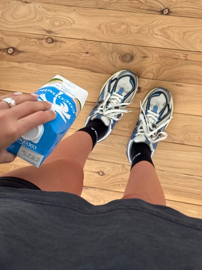 A top-down shot shows a person on a wooden floor wearing white and blue sneakers with black socks. The person is wearing a grey dress or shirt. They are holding a blue carton of "Cocobella" coconut water. The hand holding the carton has a silver ring on the ring finger. The focus is on the shoes, the carton, and the person's legs.