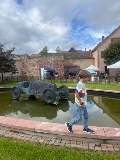 The image captures an outdoor scene in Molsheim, France, under an overcast sky. In the foreground, a young boy with light brown hair, wearing a multi-colored t-shirt, light blue jeans, and dark athletic shoes with lime green accents, walks from right to left along the red-brown stone rim of a shallow water feature containing murky green water. To the boy's left, a dark, weathered bronze statue of a vintage open-wheel racing car, resembling a Bugatti Type 35, is partially submerged in the water.

Behind the water feature, a grassy area is bordered by a low dark fence. A tall, reddish-brown stone wall, indicative of historic architecture, extends across the midground. Beyond this wall, a public gathering is evident, featuring several temporary event tents: a dark blue tent, a black tent, and two white tents. Multiple individuals are discernible under and around these tents. In the background, traditional European buildings with pitched, tiled roofs and light-colored facades are visible, including a light beige/pink building on the far right. Cobblestone paving lines the base of the fountain's rim.