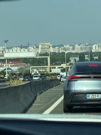 The image, taken from a vehicle, shows a city skyline in the background, characterized by densely packed buildings and some trees. A highway or elevated road is in the foreground, with several cars driving on it. The vehicle from which the photo is taken has its rear license plate visible, displaying "HC-223-FL" with a French flag emblem. The day appears bright and sunny, suggesting daytime. The scene is set on a road with a concrete barrier, and the overall impression is one of urban traffic. The image is oriented vertically, with the city and sky occupying the left side and the road and cars on the right.