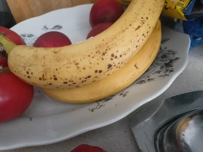 A close-up, high-angle shot captures a partially filled white ceramic platter containing several red tomatoes and two ripe bananas. The platter itself is decorated with a delicate floral pattern. The bananas are positioned diagonally across the platter, with the top banana showing a scattering of brown spots, indicating ripeness. To the right of the platter, a shiny, metallic object, possibly part of a kitchen appliance or serving dish, is visible. In the background, a portion of a yellow and blue packaged item is present. The scene appears to be set on a countertop, with a wooden surface visible at the top left of the frame. The image does not contain any elements that would allow for determining its specific location in Grudziądz, Poland.