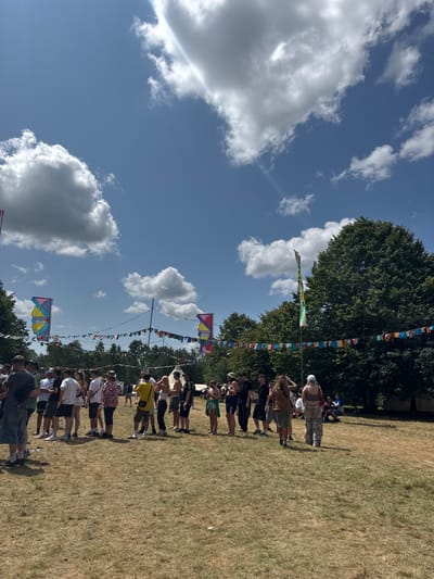 A grassy field under a blue sky with scattered white clouds is dotted with trees. A line of people stands along the field, with a string of colorful flags hanging above them. The scene captures an outdoor event or festival, with the flags adding a festive touch. The sunny weather and the presence of a crowd suggest a lively and engaging atmosphere.