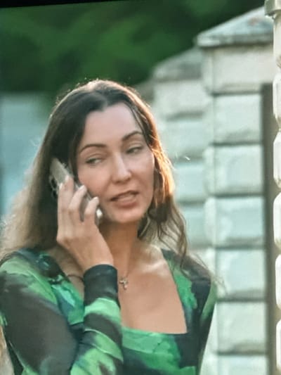 The image shows a medium shot of a woman with long, dark, wavy hair talking on a cell phone. She is wearing a green and black patterned dress with long sleeves and a square neckline. She has a small pendant necklace on. The background is blurred, but appears to be an outdoor setting with greenery and a light-colored stone structure. The scene is likely in the city of Dondușeni, Moldova, given the information provided. The woman's expression suggests she is engaged in a conversation.