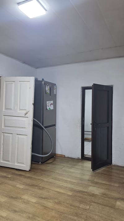 The image depicts an indoor scene, likely a kitchen or utility room, in Kochkor, Kyrgyzstan. The room features a light-colored ceiling with a modern, rectangular LED light fixture. The walls are painted white and appear to have a textured finish. On the left side of the frame, a tall, narrow, white wooden door is propped open, revealing its paneling. To the right of the door stands a modern, silver refrigerator, adorned with a few pieces of paper, possibly children's drawings. A flexible grey hose is attached to the base of the refrigerator and extends towards the floor. In the background, to the right, a dark brown door with a metal handle is partially ajar, leading to another room or area. The floor is covered with light brown wood-look laminate flooring. The overall impression is of a functional, somewhat sparsely decorated space.