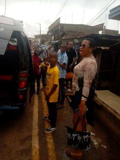 The image shows a group of people standing on a street in Umuokpu, Nigeria. A black van is parked on the left side of the frame, with several children standing nearby. A woman wearing sunglasses, a patterned top, and dark bottoms stands to the right of the children. She is holding a black handbag and appears to be speaking. The street is narrow and the buildings are densely packed. Overhead wires are visible against a cloudy sky. The street appears to be marked with painted lines.