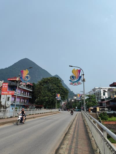 The image captures a street scene in Thành phố Hà Giang, Vietnam. The road stretches into the distance, with a person on a motorcycle riding towards the camera on the left side of the road. Another motorcycle is further down the road. The sides of the bridge are lined with metal railings, and a brick walkway runs along the right side.
On either side of the road, buildings are visible, some with vibrant signage and multiple stories. The signs are a mix of Vietnamese and other languages, including Hebrew. One sign reads "MÓN ĂN DO THÁI" and "PIZZA - MỲ Ý," suggesting a restaurant. Decorative street lamps with colorful emblems line the road.
In the background, a lush green mountain dominates the landscape, rising sharply against the clear sky. The sky is a pale blue, indicating fair weather. The overall scene suggests an urban setting nestled in a mountainous region.