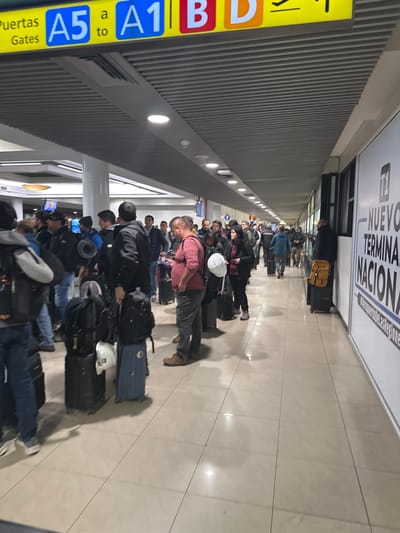 The image shows a long queue of people with luggage inside the "Nuevo Terminal Nacional" of an airport. The signage indicates gates A1 to A5 in Spanish ("Puertas Gates"). The location is likely the Arturo Merino Benítez International Airport in Pudahuel, Chile. There is a sign on the wall next to the group which says, "Nuevo Terminal Nacional".
