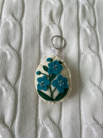 A handmade keychain with blue felt flowers is lying on a white cable knit sweater. The keychain is egg-shaped with an off-white felt background, and three blue flowers with beaded centers are sewn onto it. Green felt stems and leaves complete the floral design. A silver keyring is attached to the keychain with a short white ribbon. The sweater has a pronounced cable knit pattern with both horizontal and diagonal ridges. The photograph was taken in a place with an unknown street address in the city of Лесной Пейзаж Кп (шарапово, Russia.