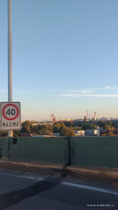 View of Dock Sud, Argentina, from a bridge. A speed limit sign indicating "40 MAXIMA" is in the foreground. Beyond that, a cityscape includes trees, buildings, and cranes. The sky is blue. The bridge's edge and road are visible to the right. Date stamp of "12 de abr. de 2026 6:02 p. m." appears vertically in the upper right.