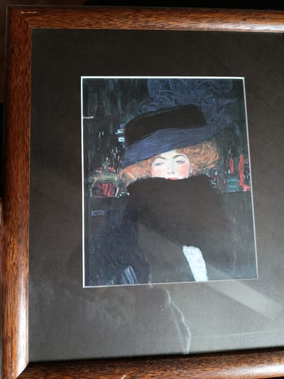 The image shows a framed painting of a woman wearing a dark hat and a fur stole. The woman has reddish-brown hair and is looking directly at the viewer with a serious expression. Her face is framed by her hat and the fur, with only her eyes, nose, and lips clearly visible. The background of the painting is dark and abstract, with hints of colors that suggest a city scene at night. A signature that reads "GUSTAV KLIMT" is visible in the lower left corner of the painting. The painting is framed with a dark brown wooden frame. The overall mood of the painting is mysterious and somewhat melancholic.