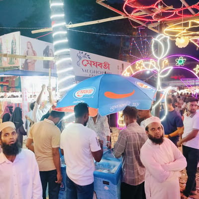 This image captures a lively night scene at a market or fair in Charfasson, Bangladesh. The setting is outdoors, bustling with numerous people, primarily men, some of whom wear traditional religious caps and beards. Women are also visible in the background, some covered with hijabs.

In the foreground, an ice cream vendor operates from a cooler, protected by a bright blue umbrella. Several men are gathered around this stall, seemingly engaging with the vendor or waiting. Decorative lights illuminate the scene, including strings of lights wrapped around a palm tree and elaborate neon light structures shaped like flowers and stars, suggesting a festive atmosphere. Large advertising banners are visible in the background, featuring images of women in fashionable attire.

The time of day is clearly night, as indicated by the dark sky and the prominence of artificial lighting. The overall mood appears active and social.

Visible text in the image includes:
*   On a banner: "মেঘবতী" (Meghoboti), "MEGHOBTI", and "FAS" (likely part of "Fashion").
*   On the blue umbrella: "সেভয়" (Savoy), "১৯৫০ সাল থেকে" (since 1950), "আইসক্রিম" (ice cream), and "ন্যাচারালি গুড" (Naturally Good).
*   On a person's shirt sleeve: "adidas".
*   In the background, on a sign partially obscured by lights: "নিজ্য মেলা/প্রদর্শন" (Nijya Mela/Prodarshan - possibly meaning local fair/exhibition).