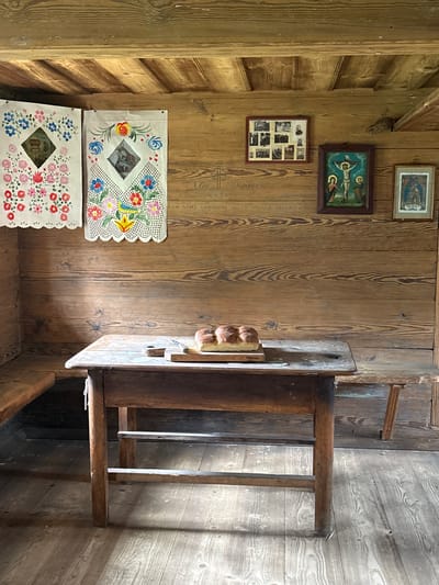 The image depicts an interior scene within a wooden structure, likely a rustic dwelling or museum exhibit, located in Sielachowskie, Poland. The walls and ceiling are constructed of aged, horizontal wooden planks, showing grain and natural wear. On the left wall, two pieces of decorative, hand-painted fabric are hung, featuring floral motifs and what appear to be religious icons in diamond-shaped inserts. To the right, a small framed collage of black and white photographs is mounted, followed by two framed religious paintings. The central painting depicts the crucifixion, with Jesus on the cross flanked by figures. The painting to its right shows a figure in blue.

In the foreground, a simple, weathered wooden table stands on a wooden floor. A cutting board with a knife and a loaf of bread, divided into four rolls, are placed on the table. To the left and right of the table, wooden benches are visible, integrated into the architecture of the room. The lighting suggests an ambient, natural light source, creating shadows on the floor and illuminating the textures of the wood. The overall impression is one of historical preservation or a staged representation of a past era.
