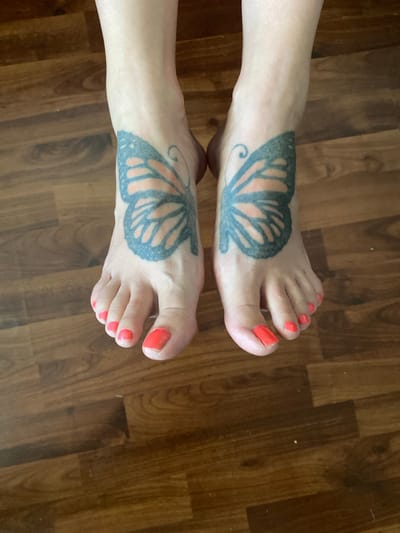 The media file features a close-up, overhead view of two human feet resting on a wooden floor. Each foot has a tattoo of a butterfly wing on the top. The butterfly tattoos are predominantly black with some orange shading. The toenails of both feet are painted a bright, coral-orange color. The lighting suggests it is daytime, and the setting appears to be indoors, likely a home. The wooden floor has a warm, brown tone with visible grain patterns. There is no discernible text in the image. The scene is static, with no activity taking place other than the resting of the feet. The overall impression is a casual and personal snapshot, highlighting body art and a bit of personal grooming.
