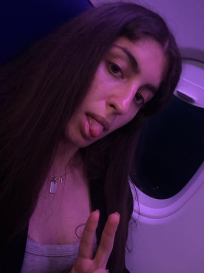 The image is a close-up selfie of a young person, likely female, with long, dark brown hair, positioned diagonally across the frame. She is seen from the chest up, with her tongue playfully sticking out and making a "peace" sign with the fingers of her left hand. She wears a silver necklace featuring a cross charm and a rectangular dog tag pendant, along with a silver ring on her left ring finger.

The setting is an airplane cabin, characterized by an airplane window visible in the upper right background, which is dark with faint distant lights, indicating nighttime. The interior cabin wall and overhead section are prominent, illuminated by a strong artificial purple light that casts a distinct hue over the entire scene, including the subject's skin and hair. A dark blue surface, likely an airplane seat, is visible in the lower left. The overall mood is casual and playful, highlighted by the subject's pose and expression. There is no visible text in the image.
