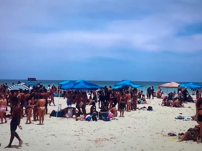 The image depicts a crowded beach scene under a clear blue sky with scattered clouds. Numerous people are scattered across the sandy beach, many in swimwear. Several beach umbrellas and tents provide shade. In the background, the ocean stretches to the horizon. The image does not provide any information that would indicate the location is Shakhovskaya, Russia.