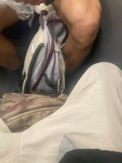 The image depicts an overhead perspective of a person's legs, covered by light-colored pants, and a backpack placed on their lap. Another person's bare leg, with visible hair, is seen in the upper right quadrant of the image. The backpack is a light beige color with maroon trim and a darker gray compartment. To the left of the backpack, a clear plastic bag filled with items is partially visible, along with what appears to be a dark blue object. The setting is indoors, with a dark gray surface forming the background and a portion of a seat visible. The image does not provide any specific visual cues that would indicate its location within the city of Sant Quirze del Vallès, Spain.