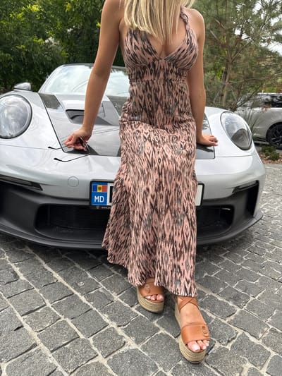 A woman with blonde hair is sitting on the hood of a silver Porsche. She is wearing a pink and brown patterned maxi dress and tan espadrille wedges. She is holding a pair of sunglasses in her left hand. The car has a license plate with "MD" on it, indicating it is registered in Moldova. The scene appears to be outdoors on a cobblestone driveway, with trees and another car visible in the background. The location is stated to be in the city of Morovaia, Moldova.