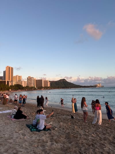 This image captures a vibrant scene at Waikiki Beach in Honolulu, United States of America. Numerous people are enjoying the beach, with some swimming in the ocean and others relaxing on the sand. The iconic Diamond Head mountain forms a prominent backdrop, and the Honolulu skyline with its tall buildings adds an urban dimension to the coastal view. The sky is clear with some light clouds, indicating a pleasant weather day. Several individuals are noticeable, including one person taking a selfie while lying on a towel, and groups of people gathered along the shoreline.