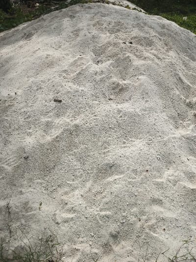 The image shows a pile of white sand. The sand is loose and granular, with a slightly rough texture. There is some sparse vegetation around the edges of the pile. The location is a pile of sand near Adanisievbe, Nigeria, which is located at coordinates latitude 5.951, longitude 5.698. No people or animals are present in the image.
