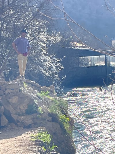 In Dračevice, Bosnia and Herzegovina, an adult individual is depicted standing on a rock formation on the left side of the frame. The person wears a light purple short-sleeved shirt, light brown or khaki trousers, a dark cap, and a bright red fanny pack worn around the waist. Their hands are placed on their hips, and a watch is visible on the left wrist. Behind the person and the rock formation, a body of water, likely a river or stream, is visible on the right, shimmering brightly due to sunlight reflection. Two dark-feathered birds, possibly ducks, are present on the water. Beyond the water, a dark-colored building with multiple windows is situated. The environment includes numerous trees, some with white blossoms or light-colored leaves, particularly behind the person, and bare branches extending into the upper right foreground. Distant hills or mountains form the backdrop. The scene is illuminated by bright sunlight.